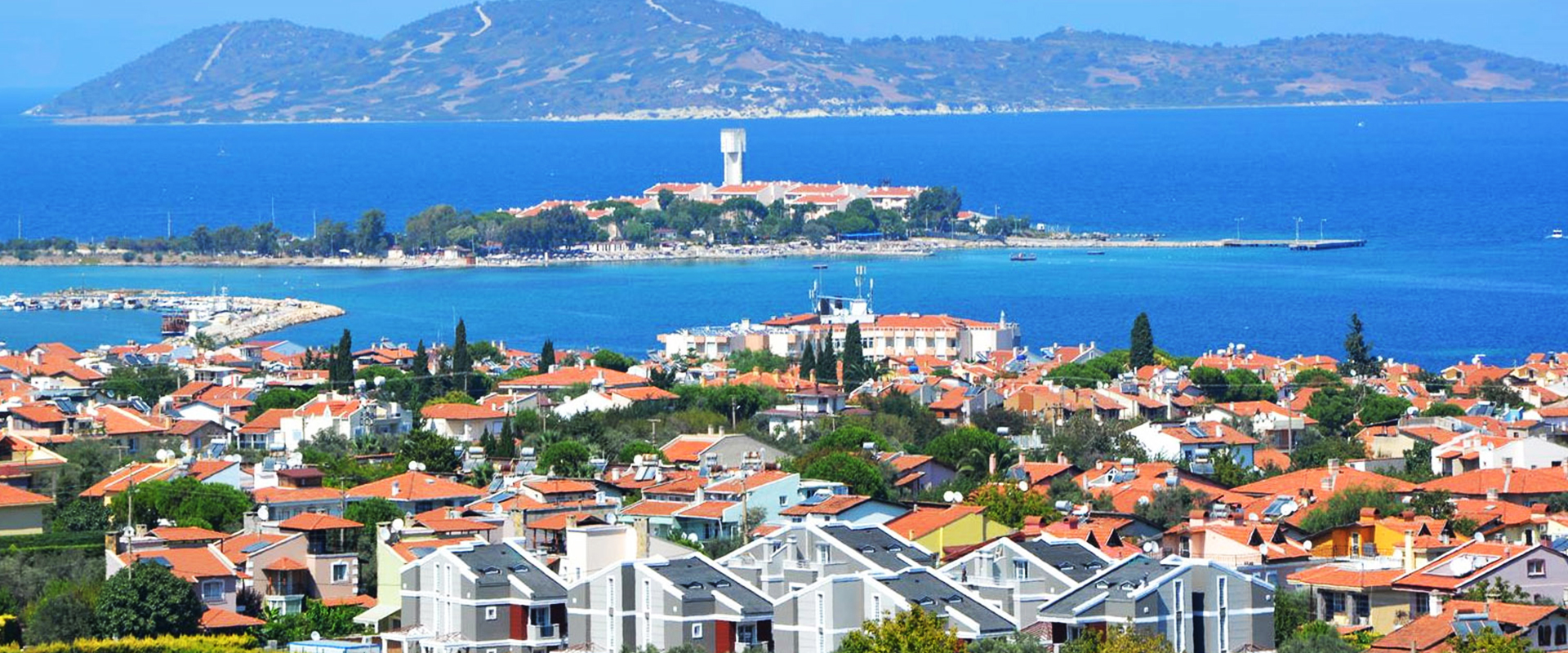 Red-roofed coastal houses overlooking a blue bay and a small island, with hills beyond.